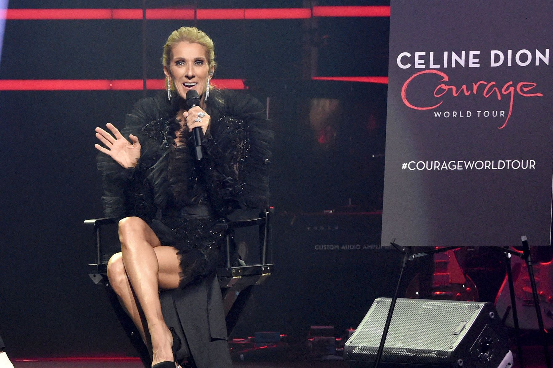 Celine Dion seated, speaking into a microphone. Black outfit, stage setting with 
