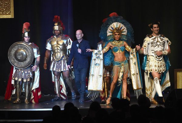 A man in black with four costumed people on a stage, two Roman soldiers, a showgirl, and a figure in a cape.