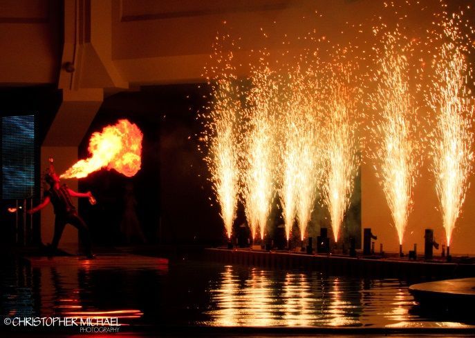 Fire performer exhales flames. Sparks erupt from fountains along a reflective water surface. Dark setting, orange light.