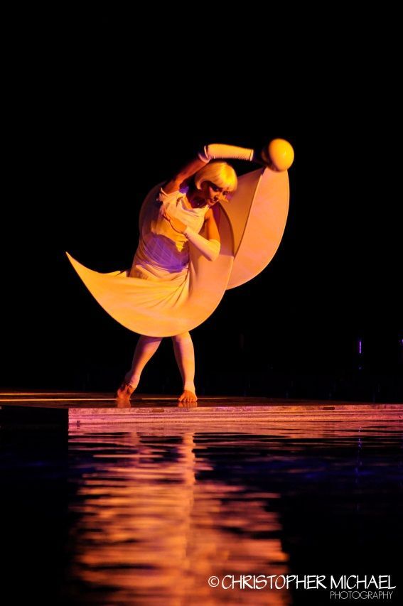 A performer in a crescent moon costume with yellow and purple details; reflecting in water against a black backdrop.