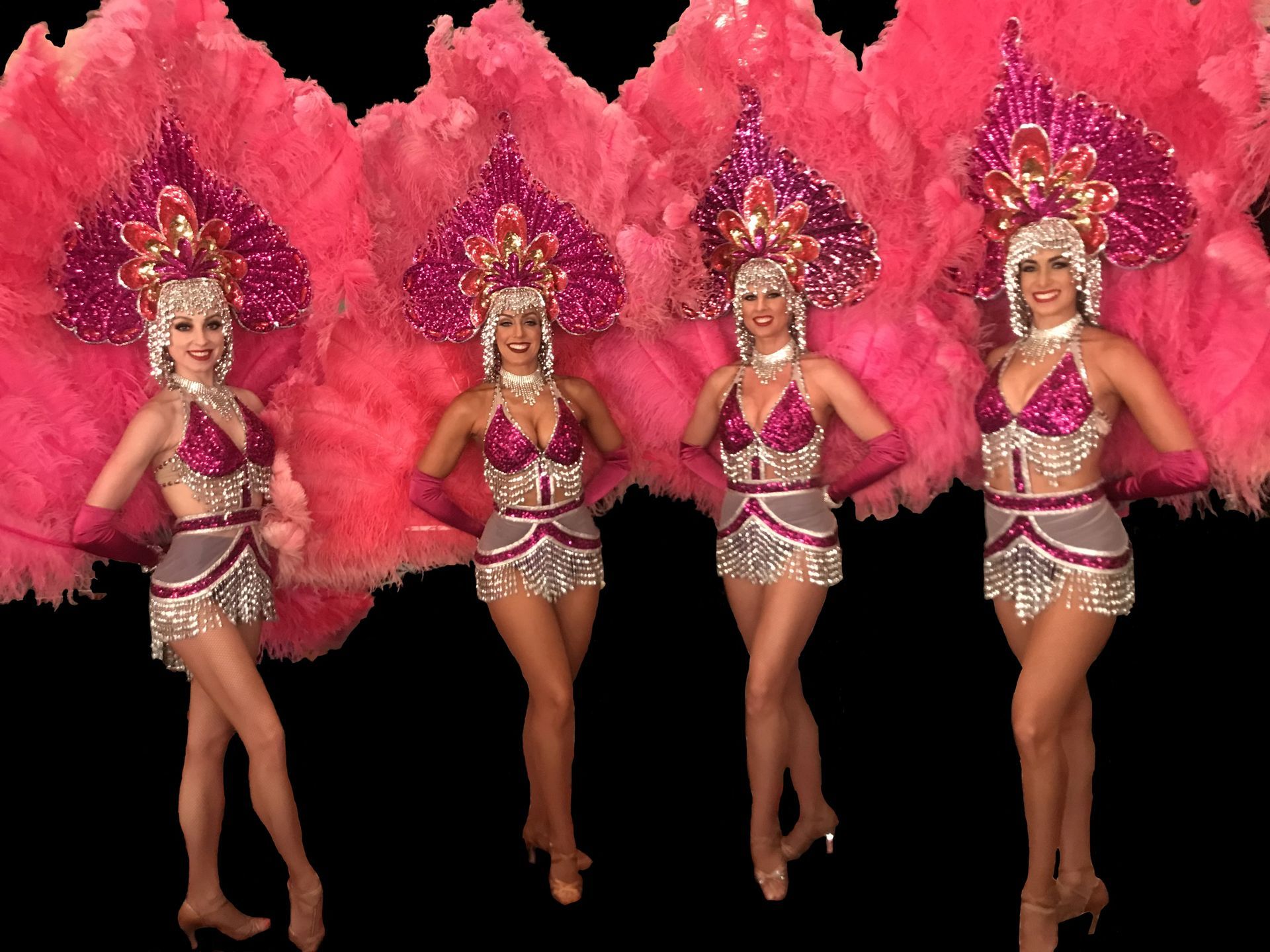 Four showgirls in pink feathered headdresses and costumes, posing against a black background.