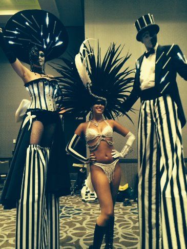 Three performers in black and white costumes. Two on stilts. A woman in the center smiles.