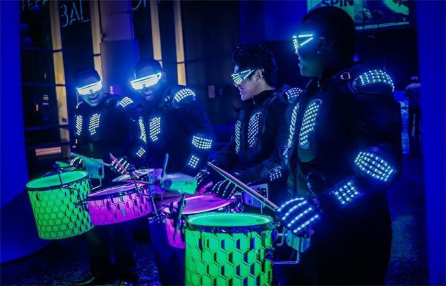 Four performers in black suits with glowing blue LEDs drumming on colorful drums in a dark setting.