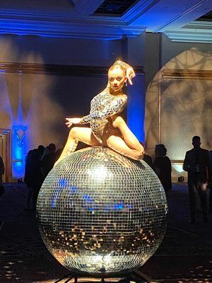 Woman in sparkly bodysuit poses on a large disco ball at an event.