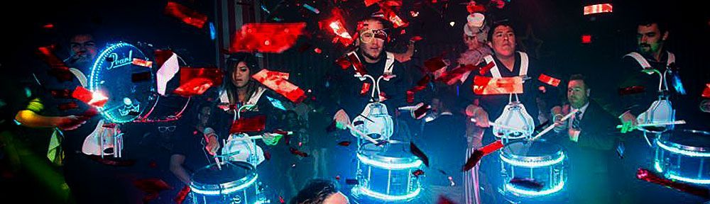A group of musicians performing in a club-like setting, confetti falling. Musicians are drumming on lit drums.