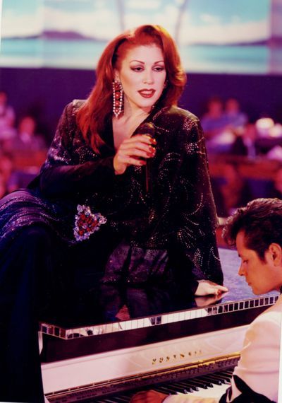 Woman with red hair singing, leaning on piano, black sequined jacket.