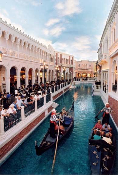 Gondolas travel a canal with people dining along a street lined with buildings in the Venetian style.