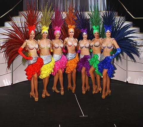 Showgirls in colorful costumes with feathered headdresses and ruffled skirts, posing on stage.
