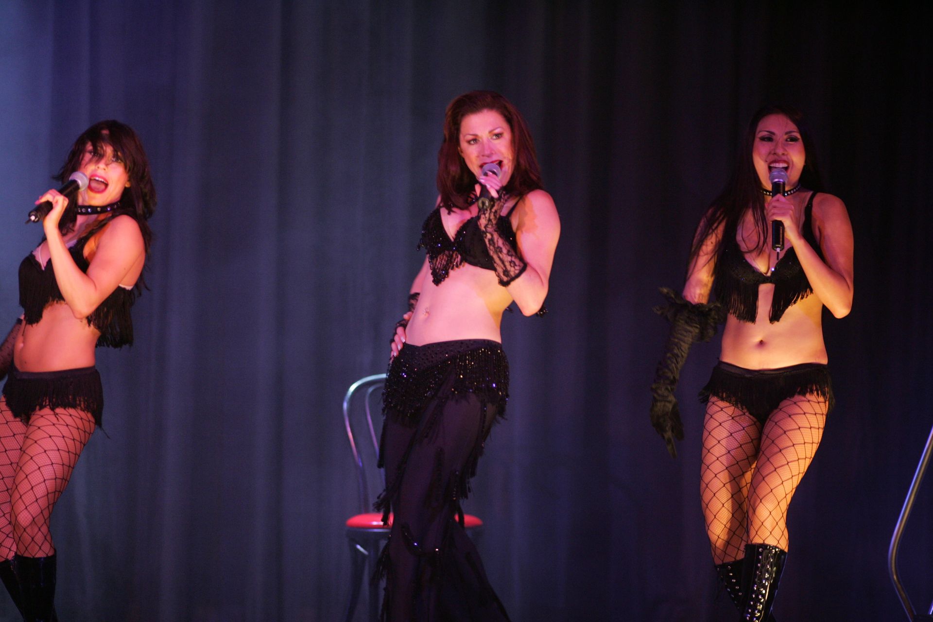 Three women in black lingerie sing on stage under blue lights.