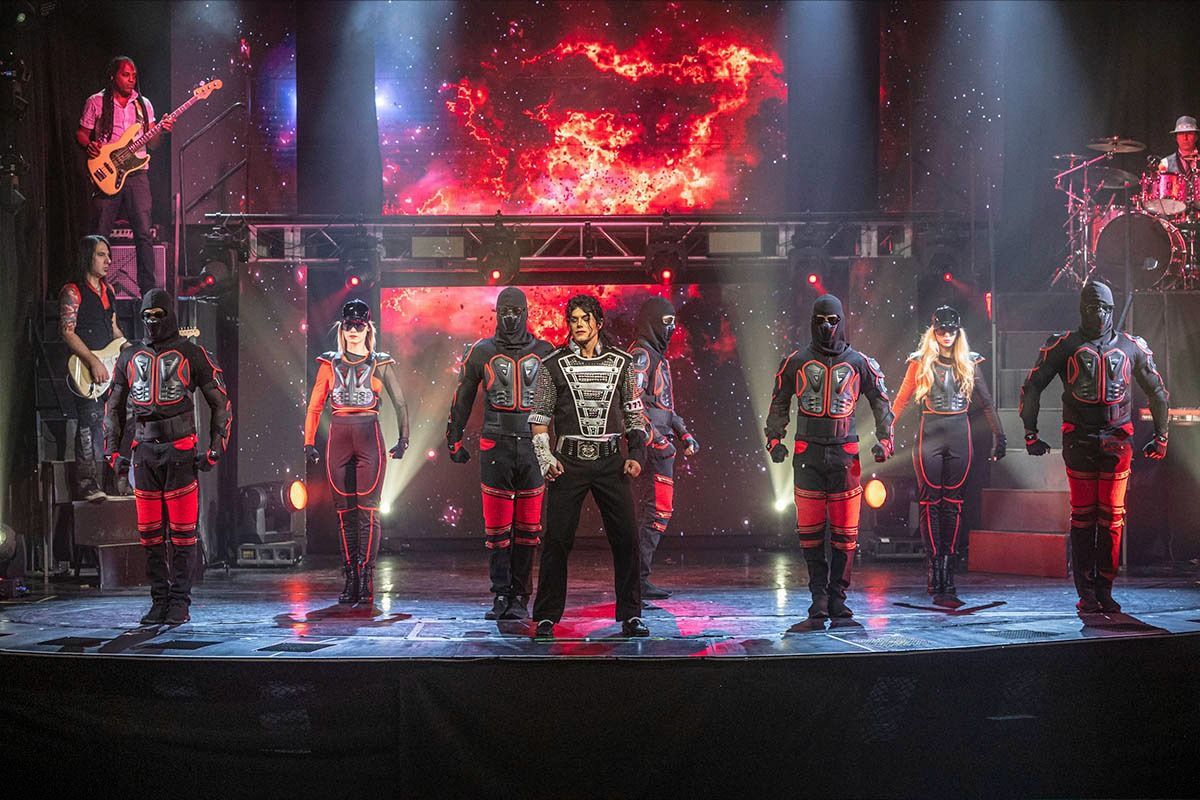 A band performs on stage. A central figure in silver stands before masked figures with red accents, amidst bright lights.