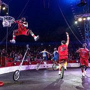 Basketball player in red leaps to dunk, circus performance with unicycles, audience in background.