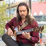 Man with long hair playing a brown guitar while seated, wearing a burgundy shirt, in front of a window.