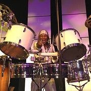 Drummer smiling, pointing, playing large white drums on stage with purple lighting.