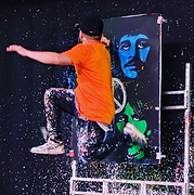 Man jumps over a canvas with colorful portraits, wearing orange shirt and paint-splattered pants.