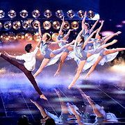 Dancers in a ballet routine on stage with bright lights. Several in blue dresses leap, one man does the splits.