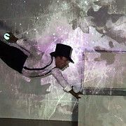 Man in hat, shirt, and dark pants reaching towards a box, with a glowing ball in hand. Against a purple and white wall.