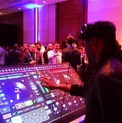 DJ at a party, mixing music on a large console. Purple and blue lighting, crowd in background.