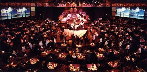A large ballroom with many people seated at tables, a stage with performers, and screens above.