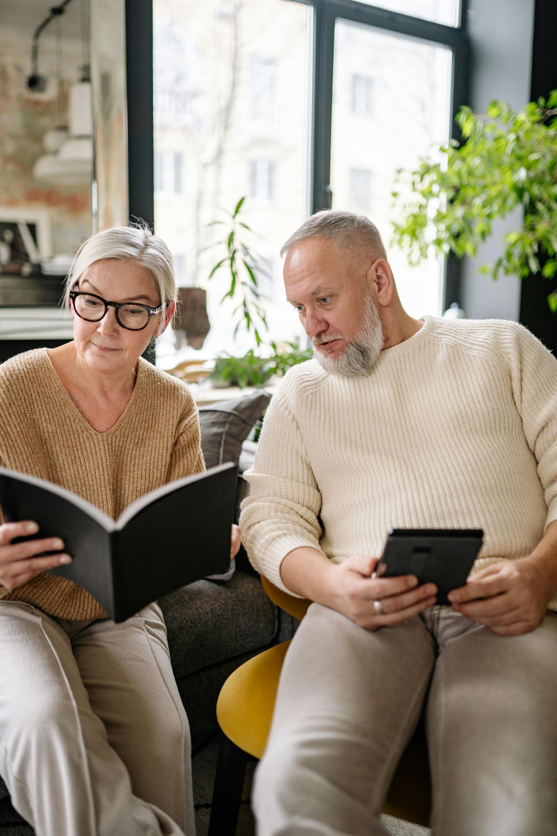 An elderly couple is sitting on a couch reading a book and using a tablet.