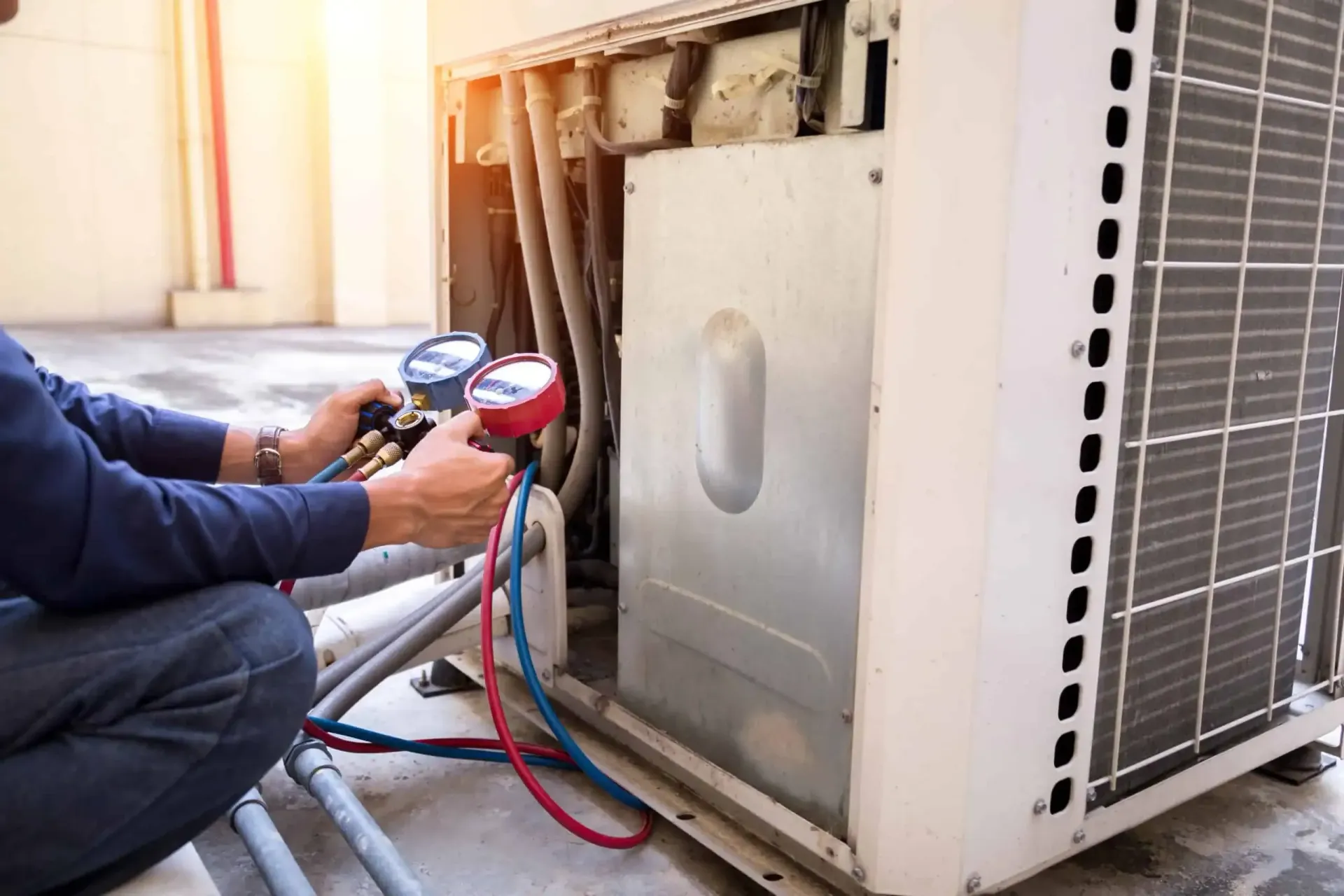 Technician servicing an AC unit, checking pressure gauges with red and blue hoses outdoors.