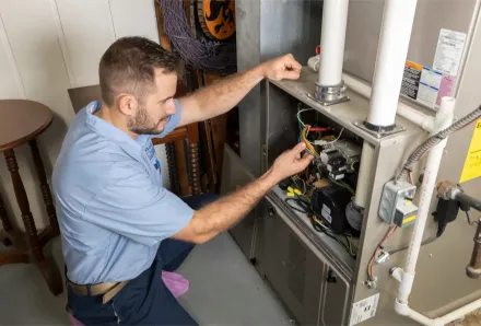 Man inspecting furnace wiring, indoors. He's crouching, wearing a blue shirt and focused.
