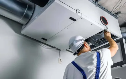 HVAC technician in white hard hat adjusts ductwork on ceiling unit. White, gray, and blue tones.