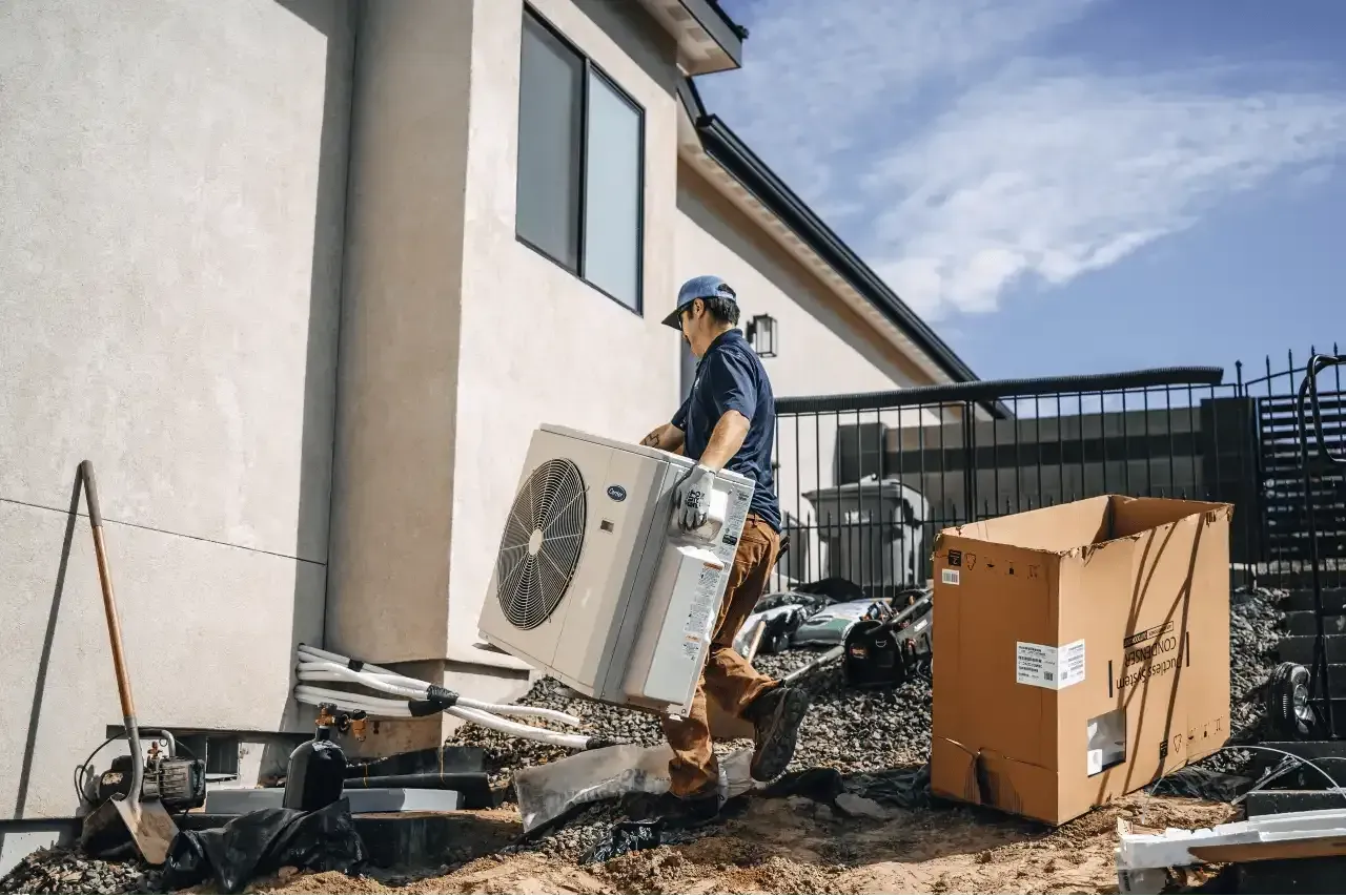 A technician carrying an air conditioning unit next to a house under a blue sky.