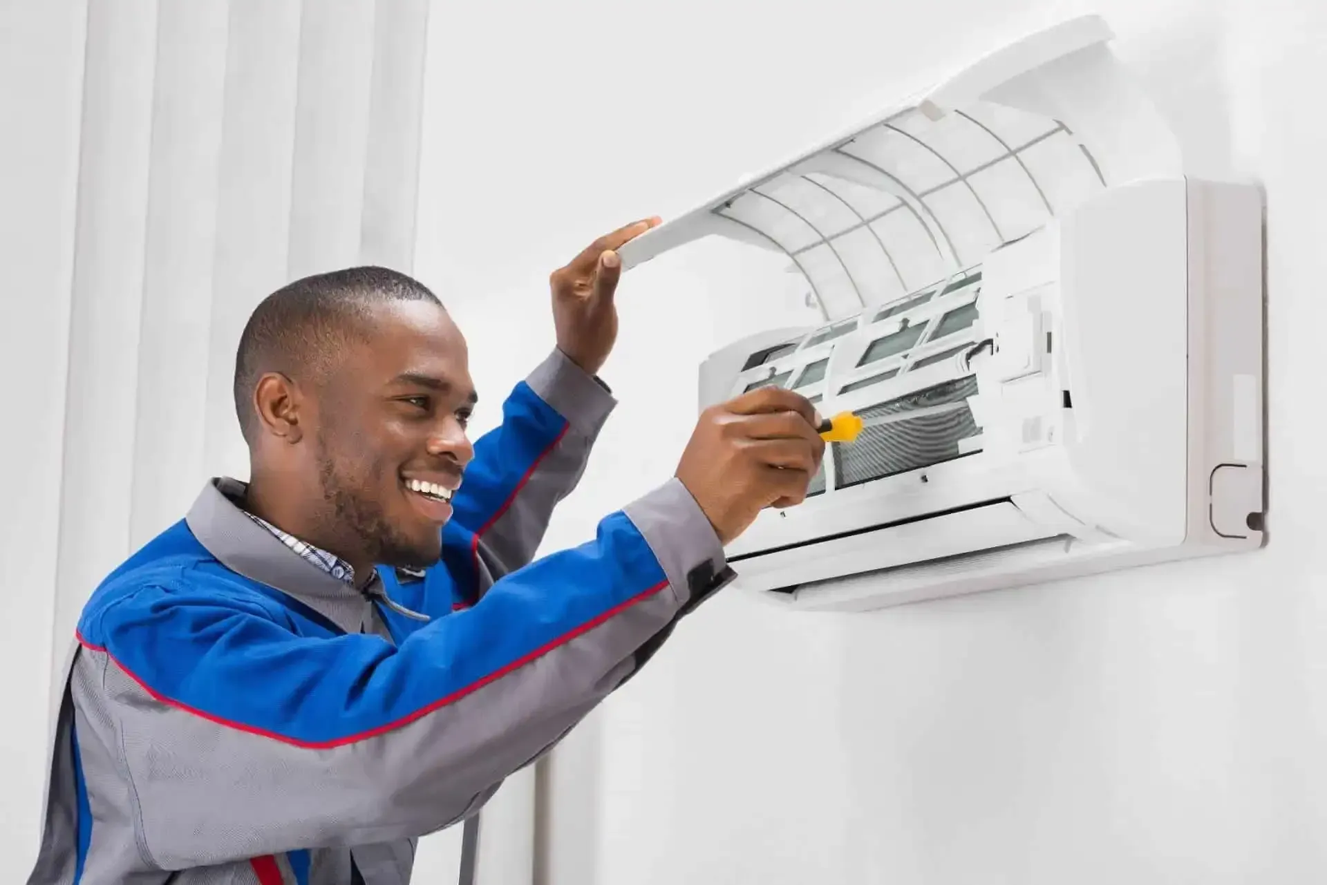 Man in blue uniform smiling while servicing a wall-mounted air conditioner with a screwdriver.
