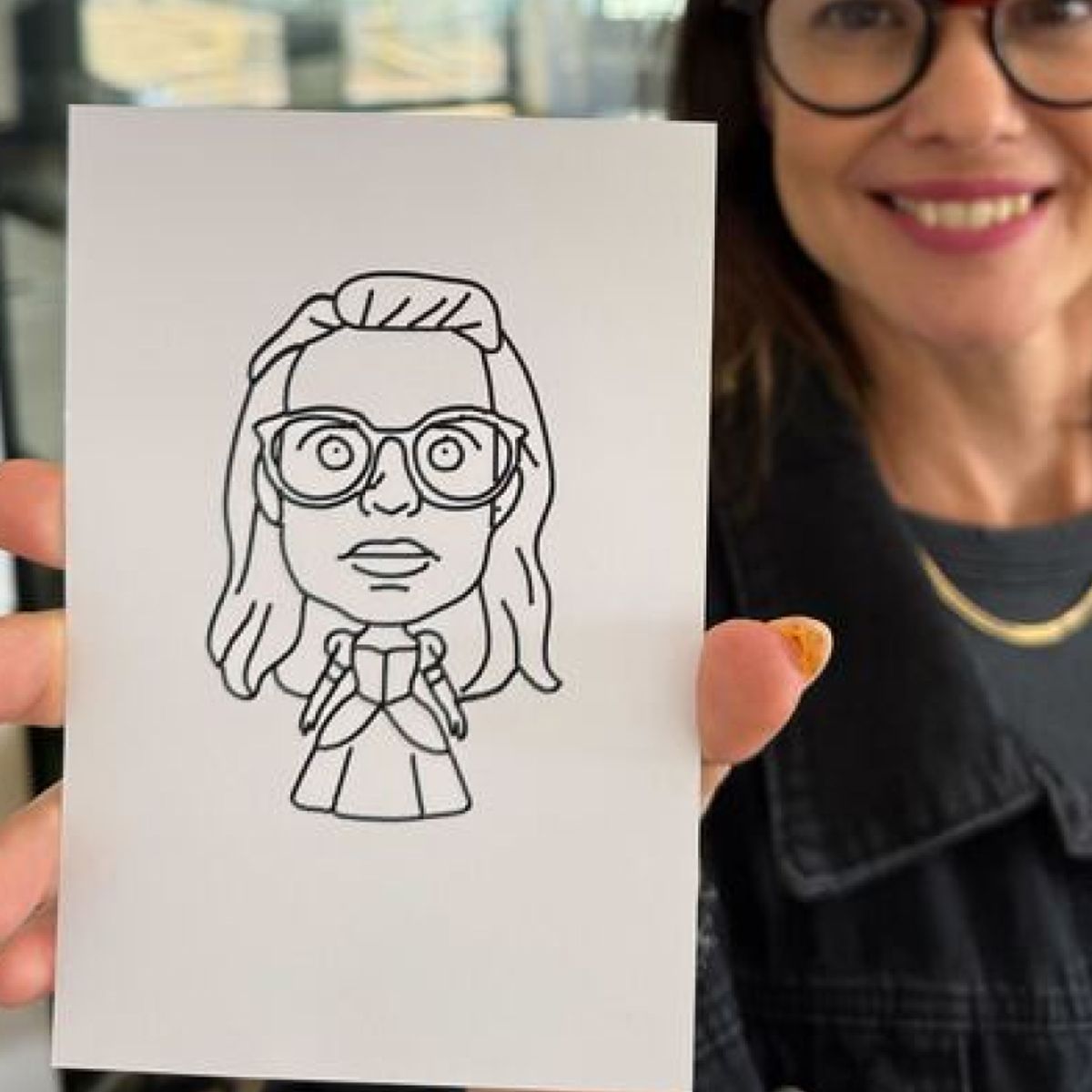 A person holds a piece of paper featuring a line-art caricature of a person wearing glasses and a dress.