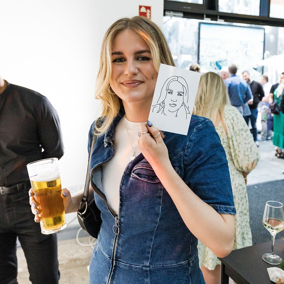 A smiling person in a denim dress holds a small hand-drawn sketch, while holding a glass of beer in the other hand.