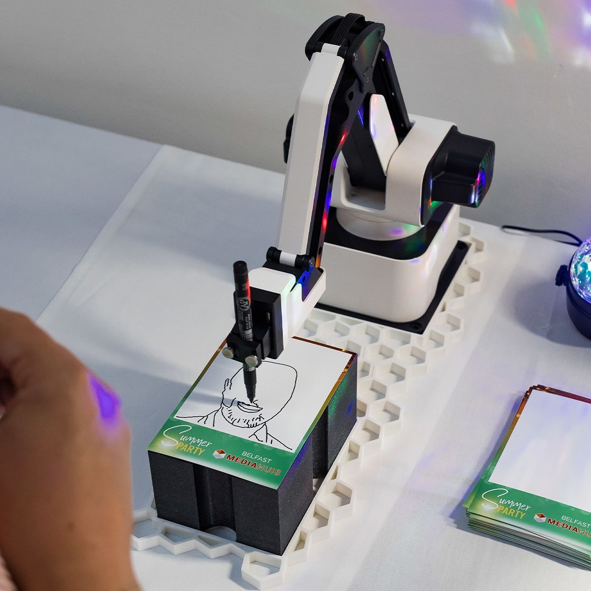 A small white robotic arm with a pen is drawing a portrait on a notepad on a white desk.