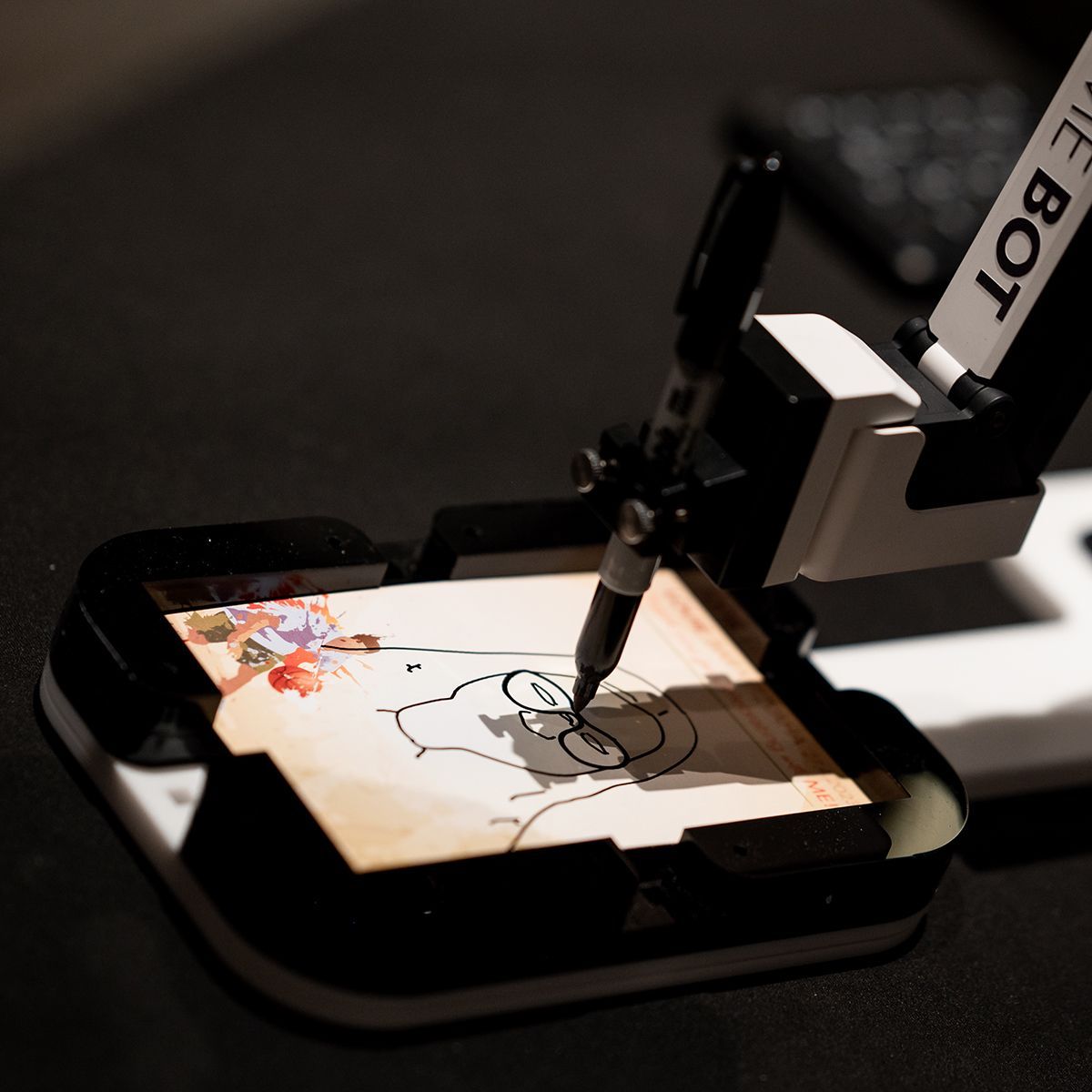 A robotic arm holds a pen, tracing the outline of Homer Simpson onto a small piece of paper held in a black frame.