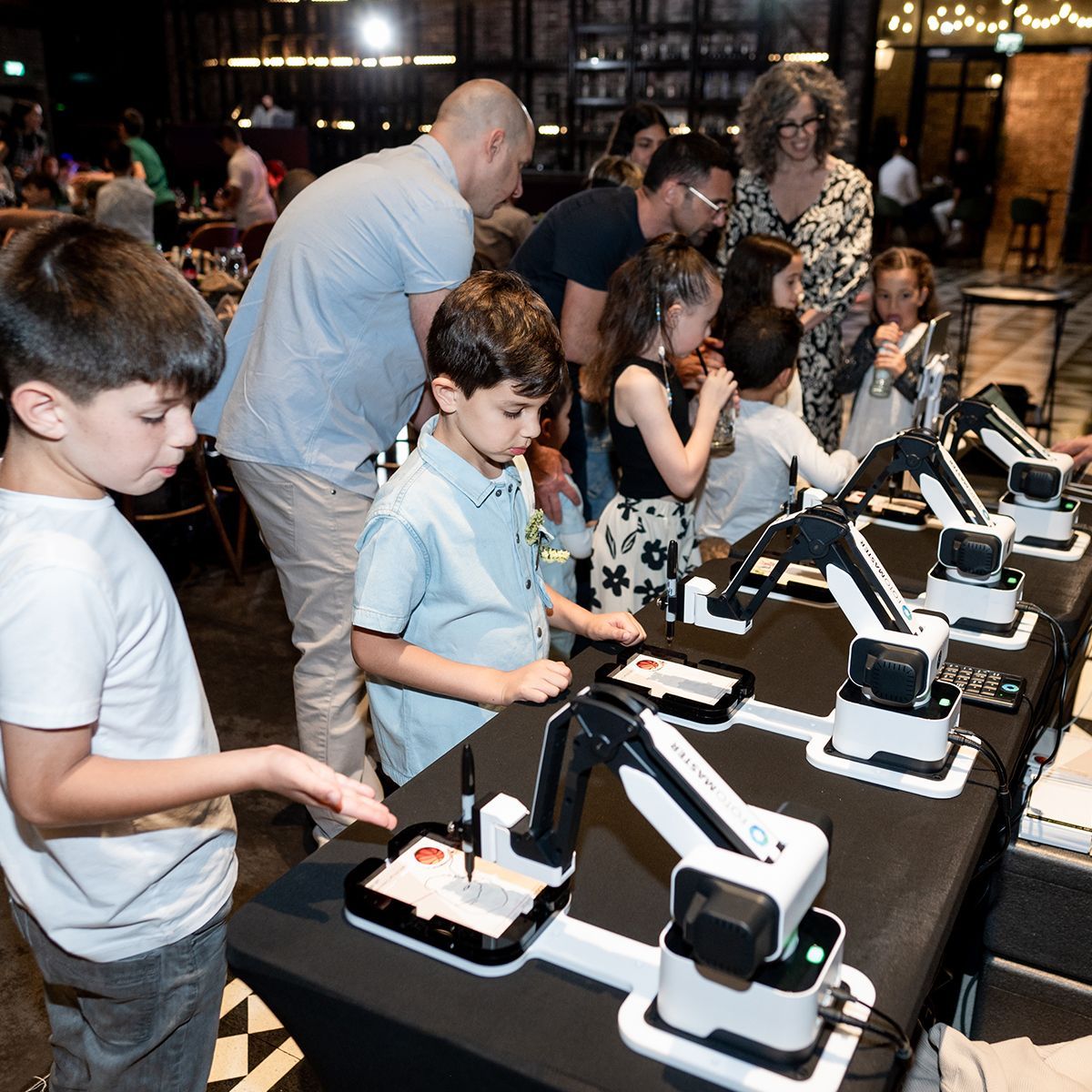 Children interact with small robotic arms on a long table in a workshop setting, watching the devices draw on paper.