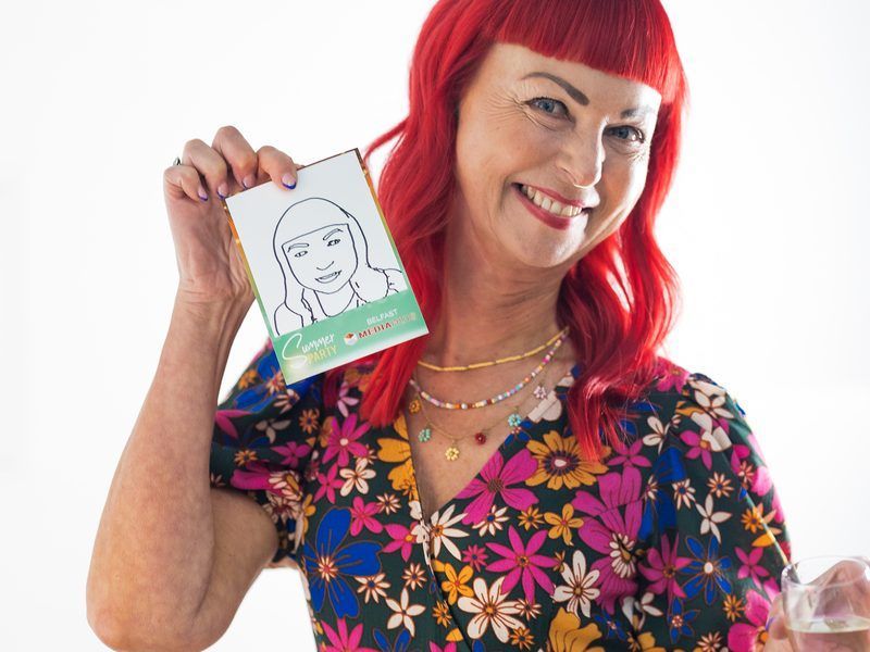 A person with vibrant red hair and a floral dress smiling while holding a small sketch of themselves against a white wall.