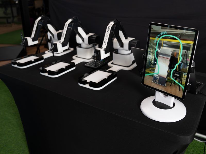 Four small, white robotic arms stand on a black table next to a tablet displaying a facial tracking overlay.
