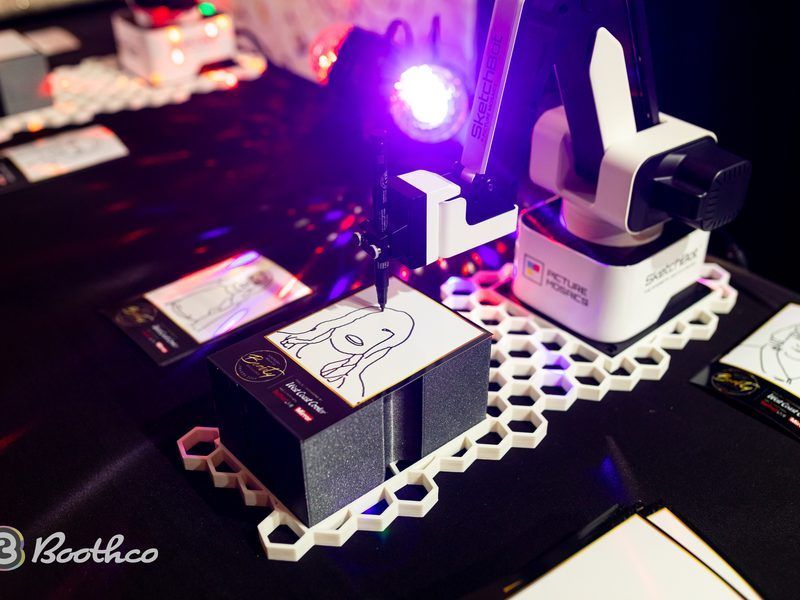 A robotic arm draws a portrait on a stack of cards resting on a white hexagonal grid platform under purple lighting.