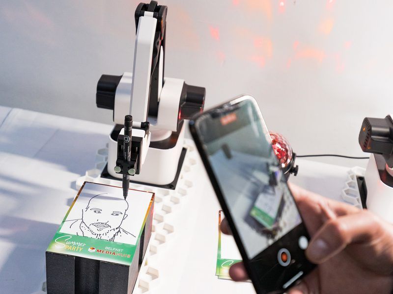 A robotic arm draws a portrait on a notepad while someone records the process with a smartphone.