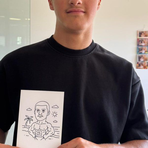 A person in a black shirt holds up a small, hand-drawn illustration of a muscular character on a beach with a palm tree.