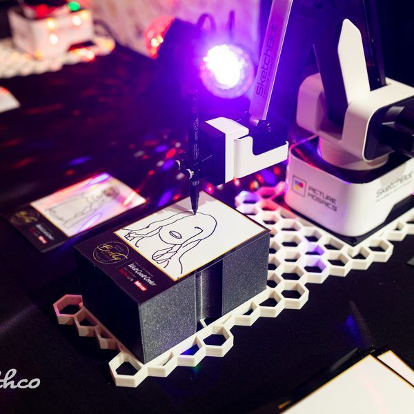 A robotic arm draws a portrait sketch on a small card resting on a white hexagonal grid platform under purple lighting.