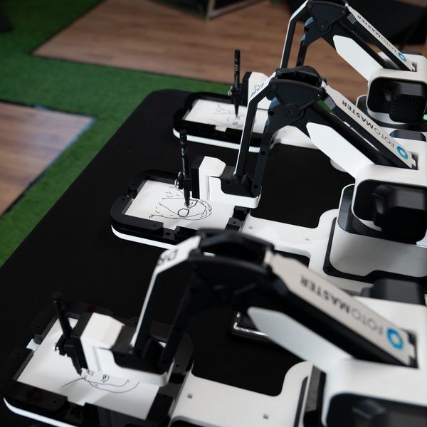 Three white-and-black robotic arms draw sketches on paper surfaces arranged on a black table.
