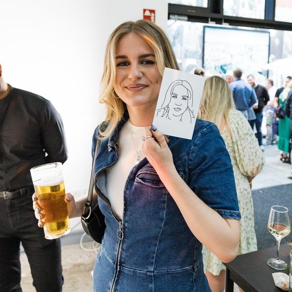 A smiling person in a denim dress holds a small sketch of their face while holding a glass of beer at an indoor event.