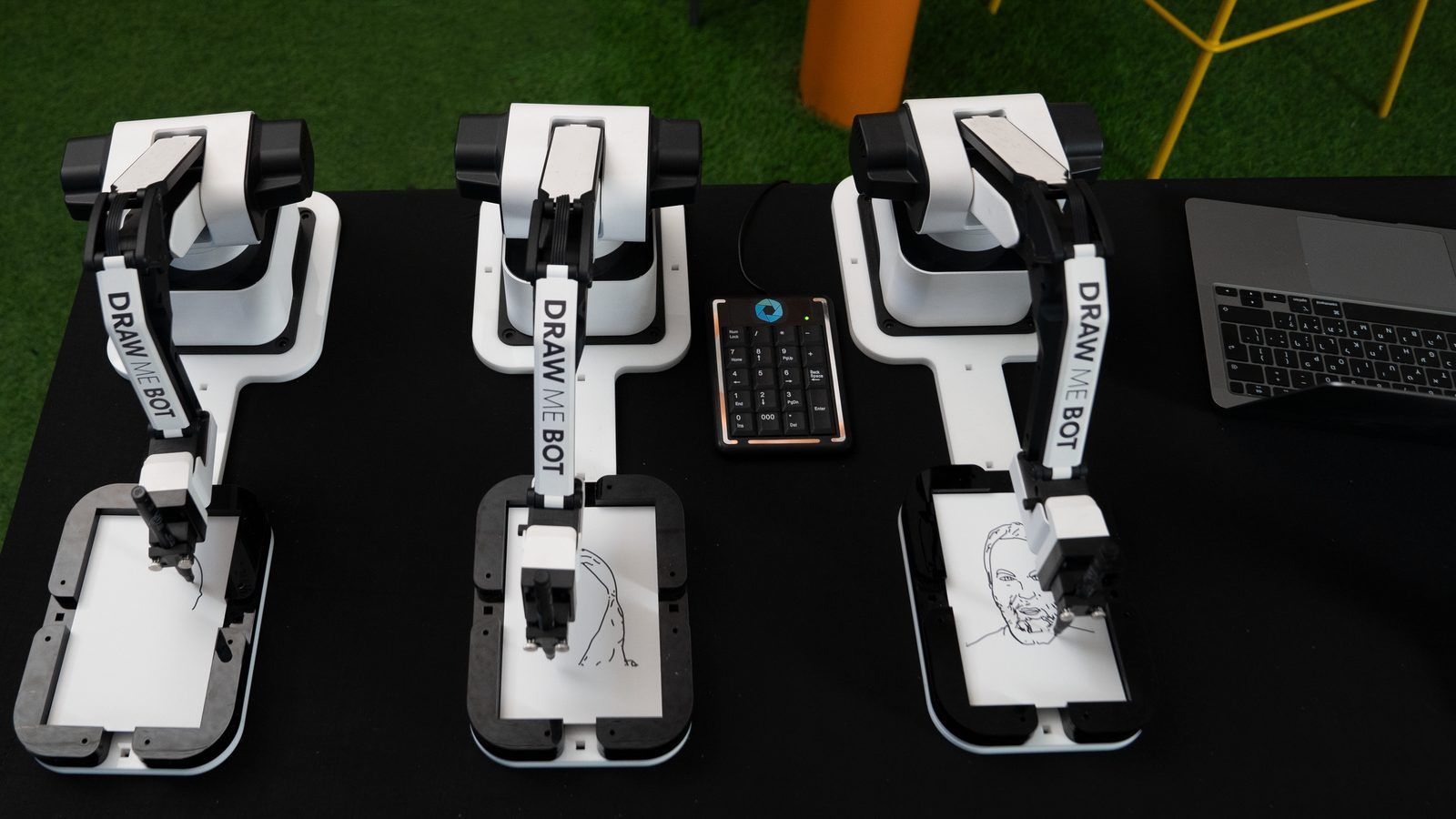 Three white robotic arms on a black table, each drawing portraits on small white pads with a control pad in the center.