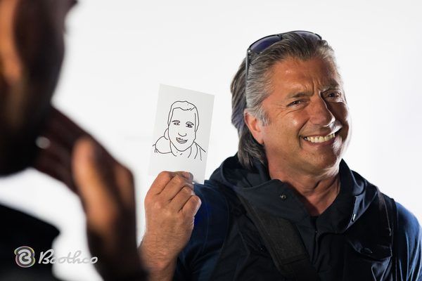 A smiling person holds up a hand-drawn sketch of a face, with a blurred person in the foreground.