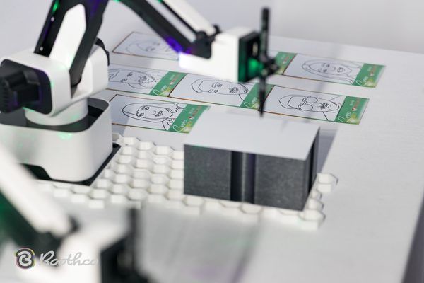 A robotic arm uses a pen to draw portraits on rectangular cards arranged on a white work surface.