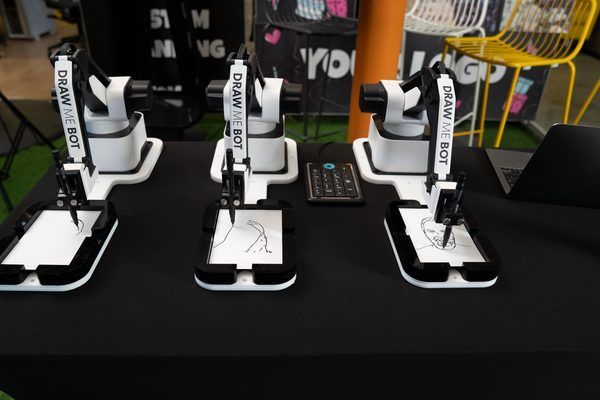 Three robotic drawing arms sit on a black table, each sketching on a small white sheet of paper.