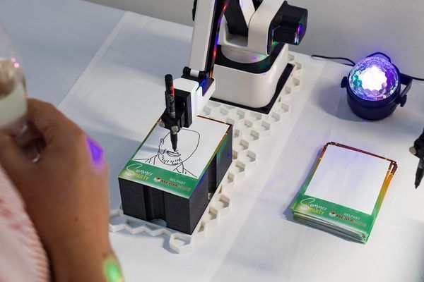 A robotic arm draws a portrait on a notepad on a white table next to a small light fixture.