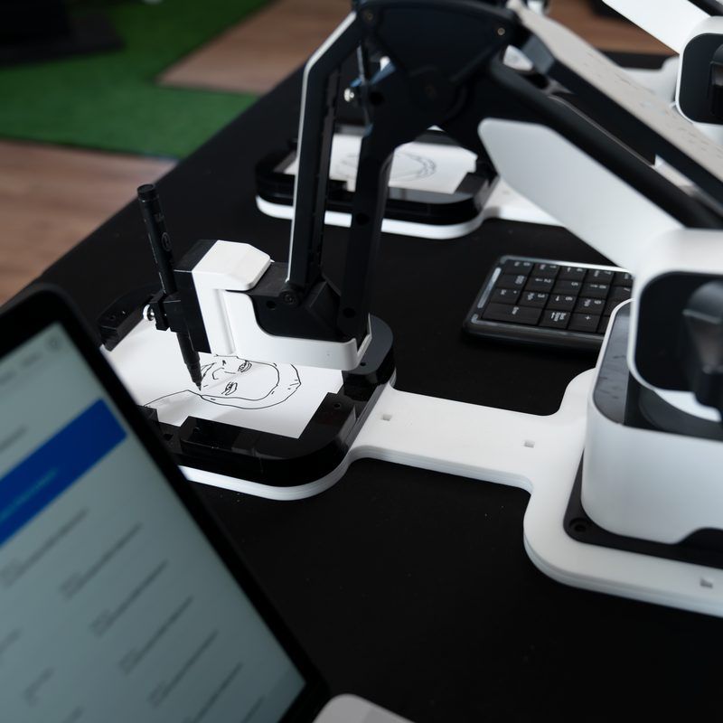 A robotic arm draws a portrait on paper, controlled by a laptop computer in the foreground.