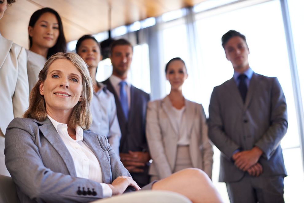 A smiling businesswoman in a gray suit sits in front of a diverse team of colleagues, all in business attire.