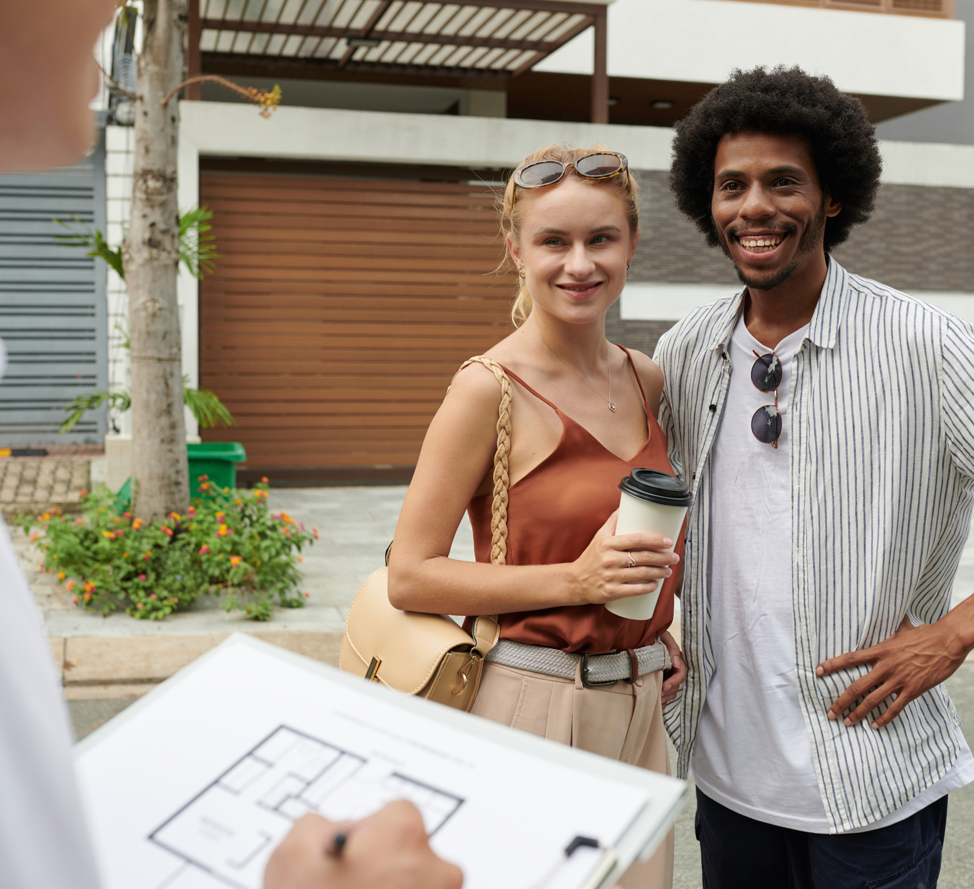 Couple smiling outdoors while looking at a home plan with a person holding the clipboard in front of them.