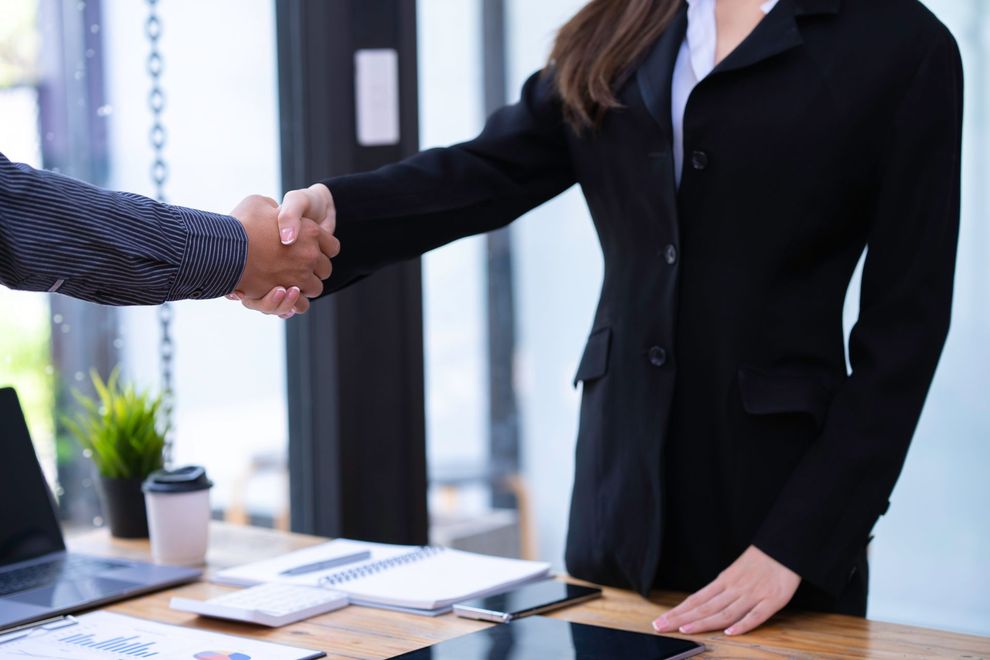 Two people shaking hands across a wooden desk in an office setting, one wearing a black blazer, the other in a patterned shirt.
